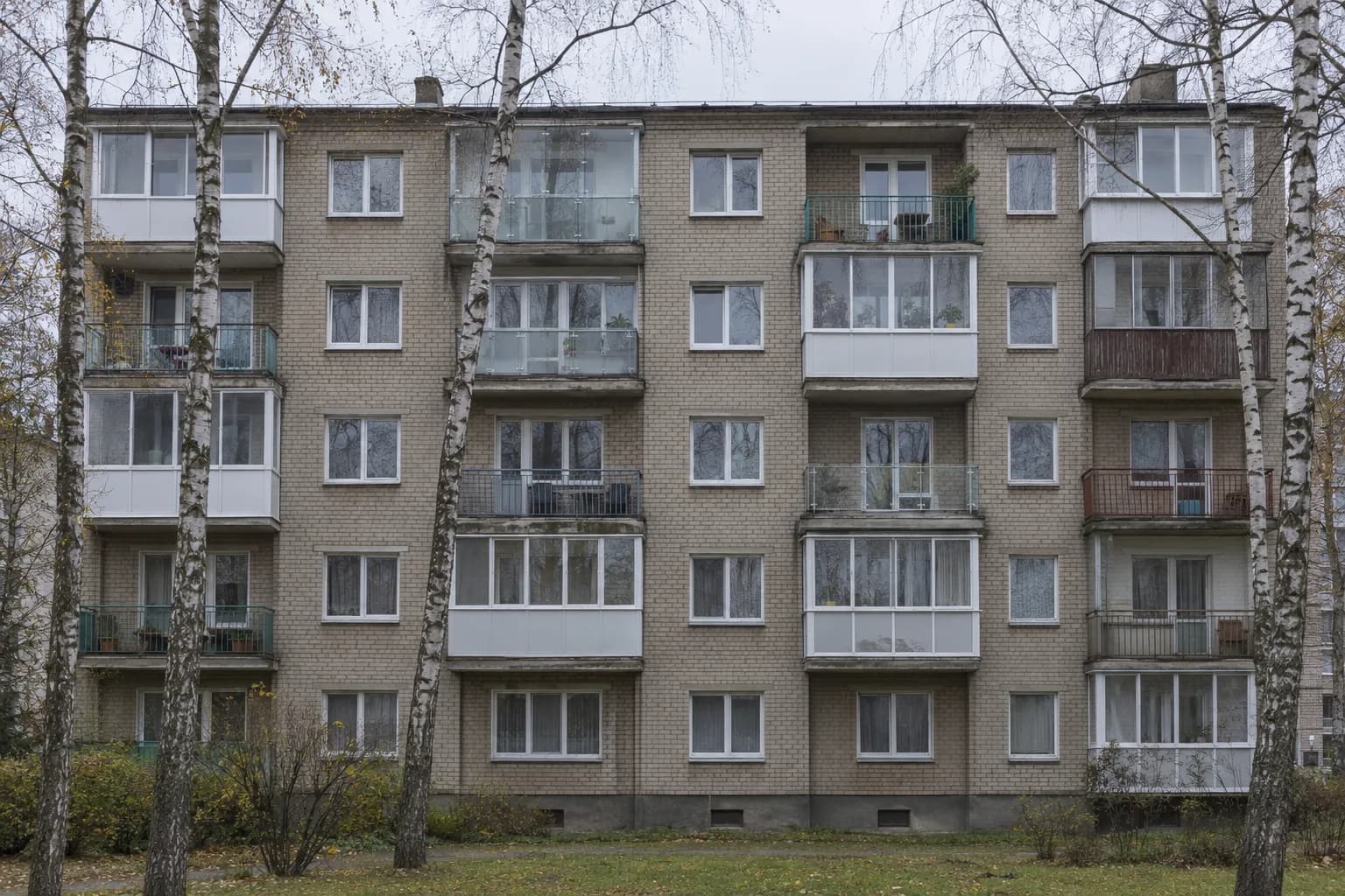 Sovietmečio penkiaaukščio plytų daugiabučio fasadas: dalis balkonų įstiklinta baltais rėmais ar bekadriu stiklu, dalis dar atvira su metaliniais turėklais